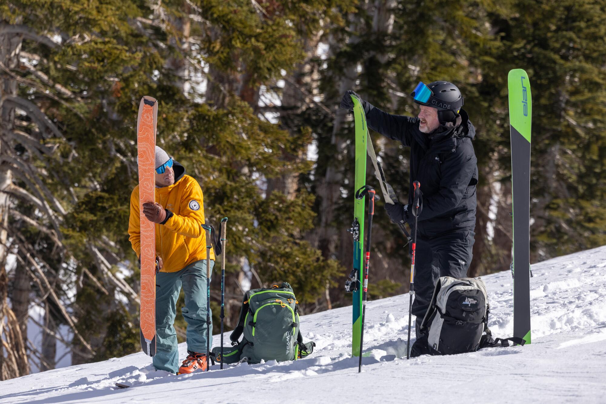 Two skiers pause on a snowy slope to install climbing skins on their backcountry skis.